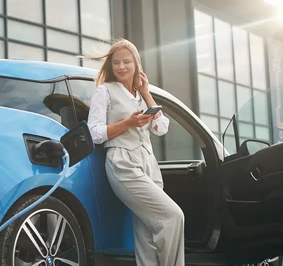 Woman holding a phone leaning against her green electric car as she looks up the Jade Finance car loan balloon calculator.