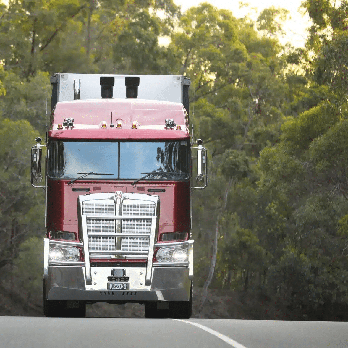 A dark red big rig is heading straight toward the camera on a road with trees in the background