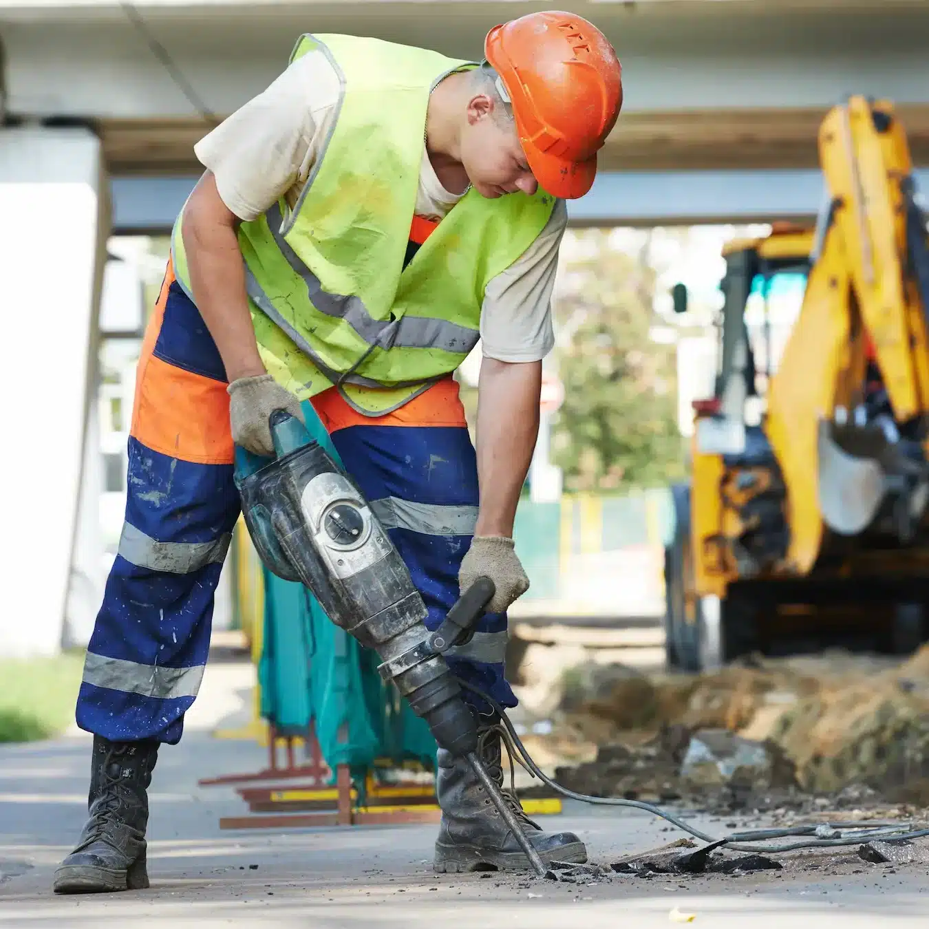 A construction working using a jack hammer to break up a concrete slab with a backhoe machine in the background
