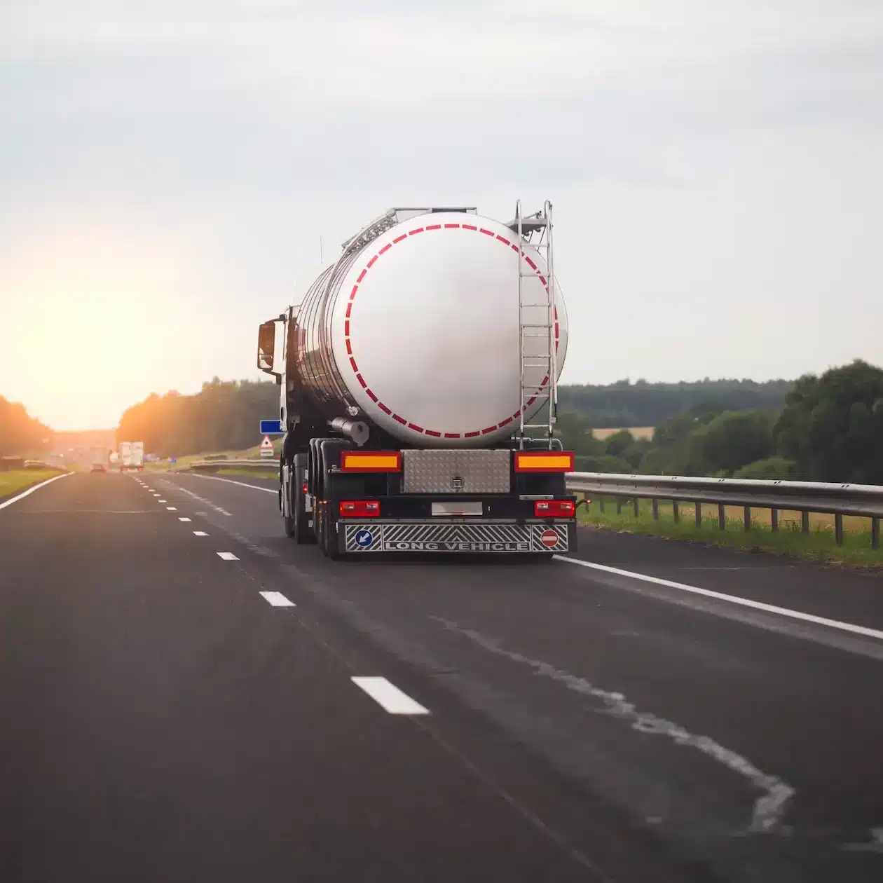 A tanker truck driving away on a long straight road towards the setting sun