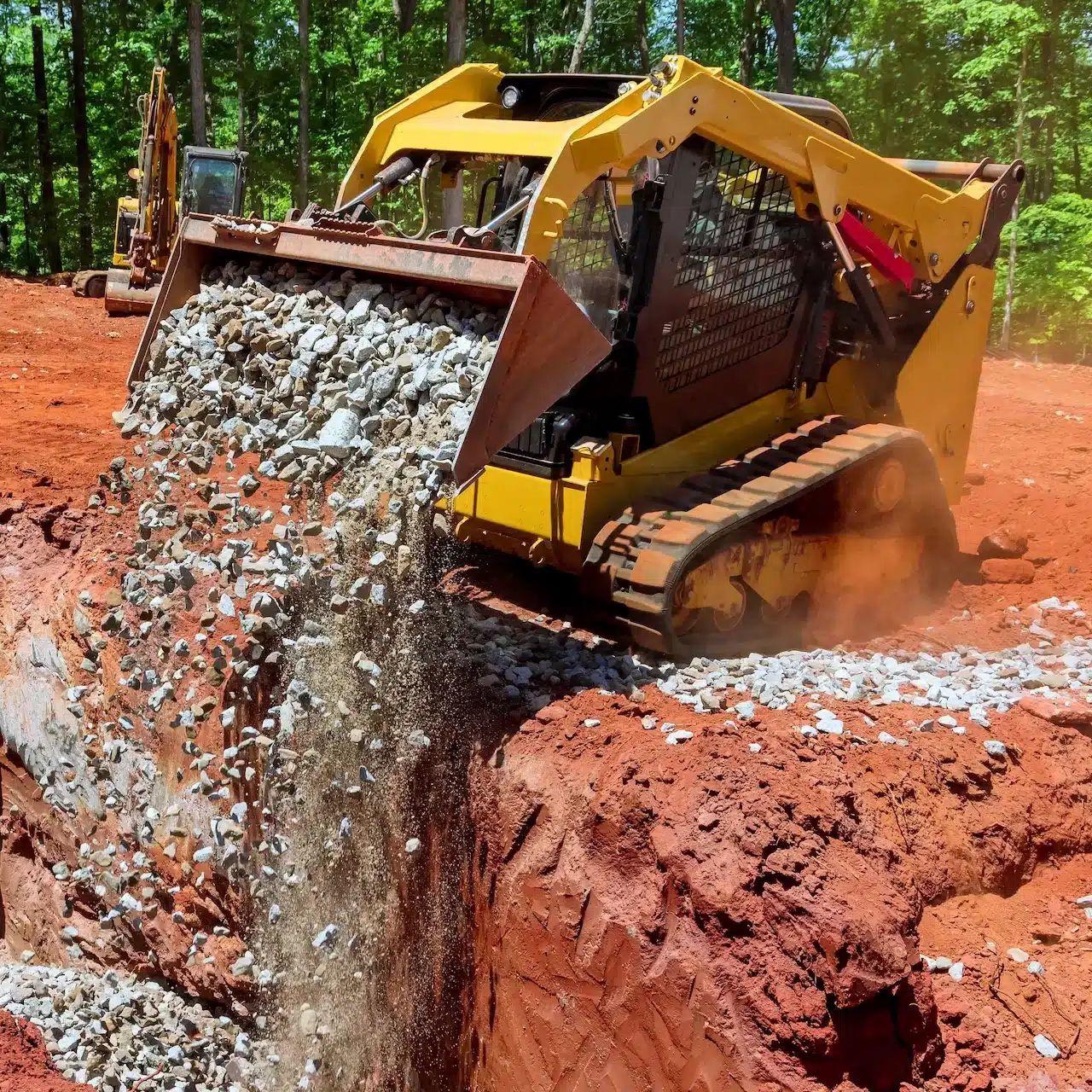 A skit steer loader is tipping it's bucket full of crushed rock into a trench dug in the ground