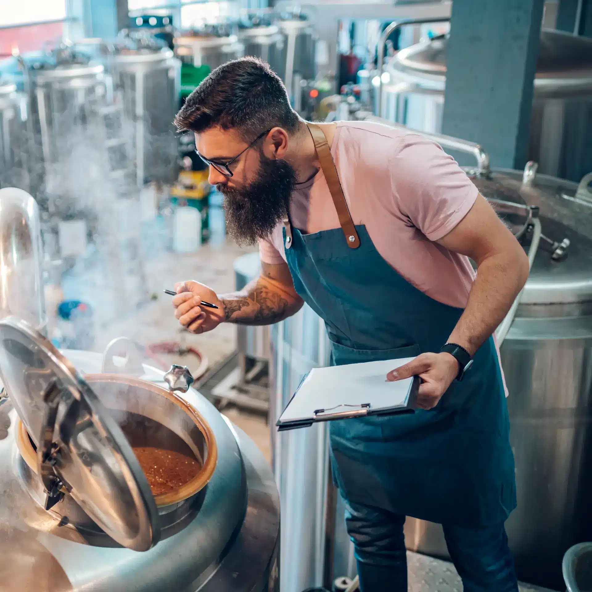 Man wearing an apron holding a clipboard and pen leaning over an open vat checking the beer brewing process.