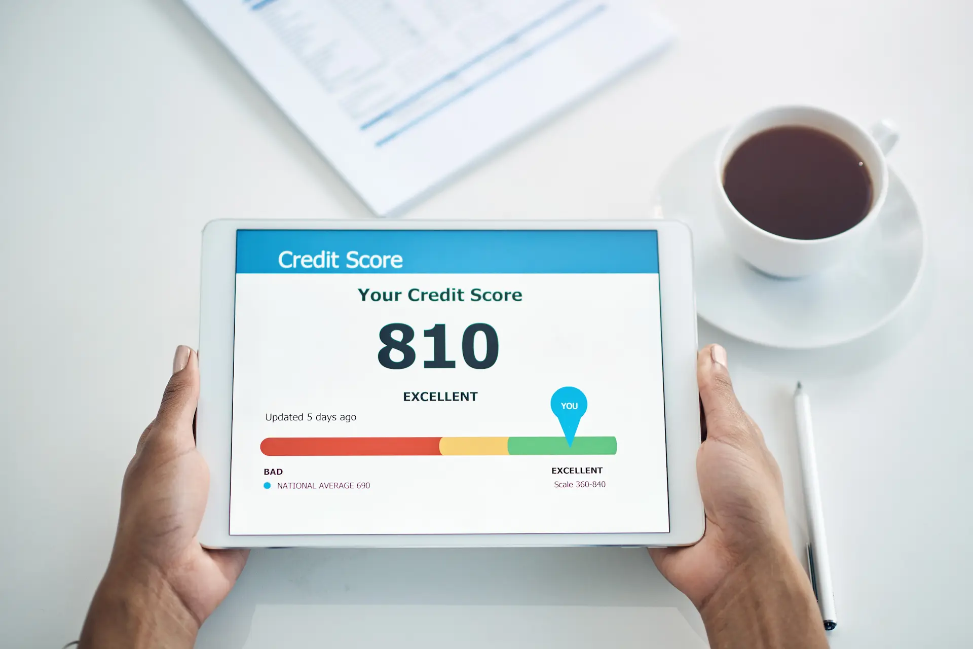 Person holding their tablet with credit score displayed on the screen, held just above white table with paper and coffee around it