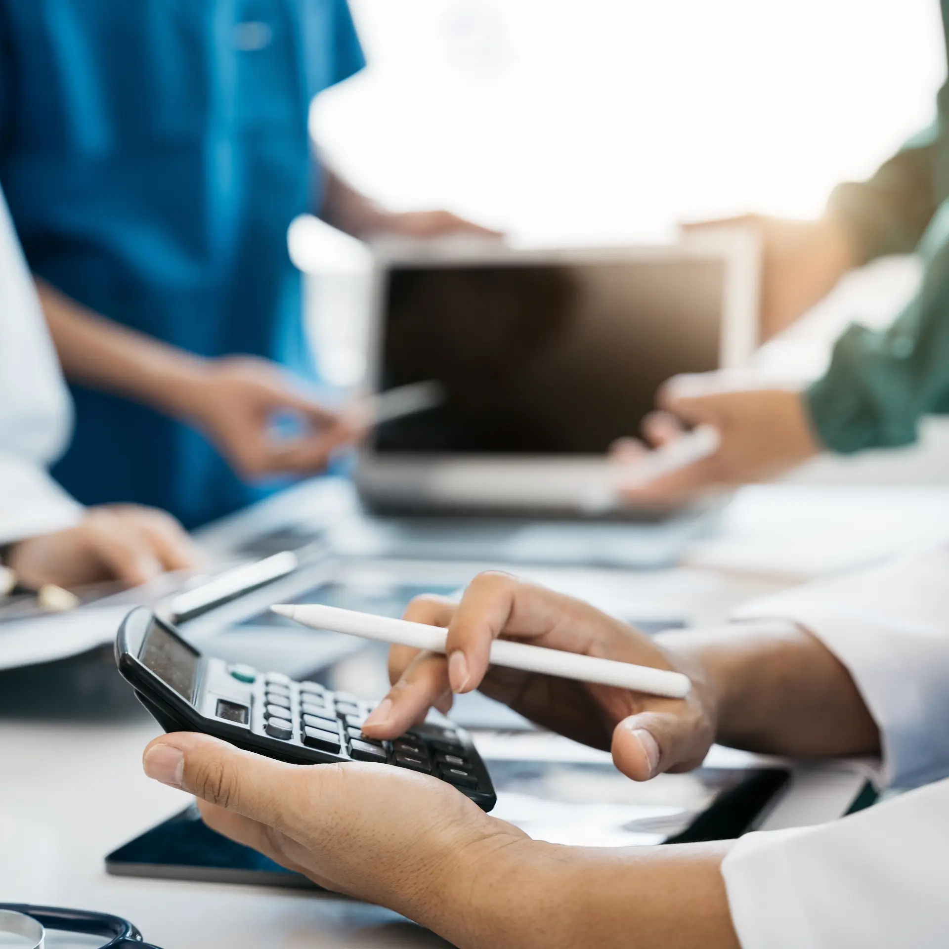 A doctor is using a calculator and a pen at a desk to work out a medical bill. Others in the background are engaged with a laptop and documents in a medical setting.