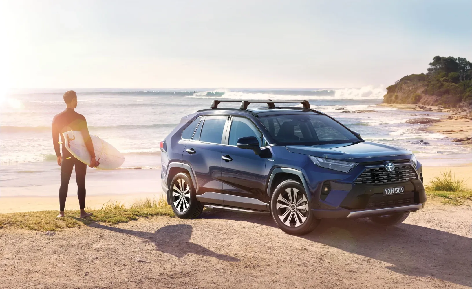 A surfer with his surf board is standing next to a Toyota RAV 4 car looking out over the ocean from the beach