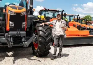 A man is standing in the dealer yard of a tractor manufacturer holding a brochure in one hand, with his other hand on the front tyre of a large new orange tractor