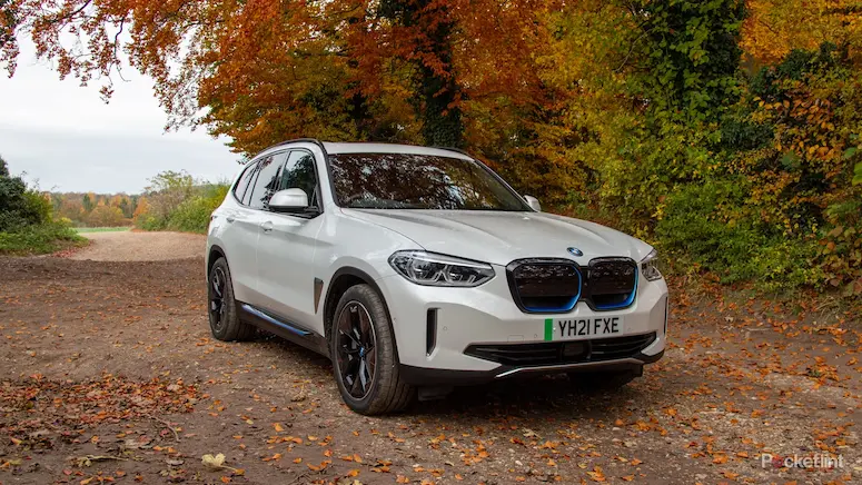 White BMW iX3 electric car parked on a dirt path with autumn trees in the background