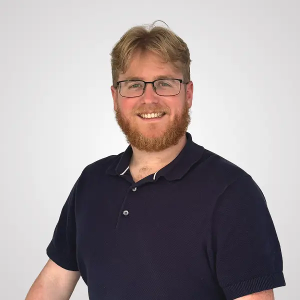 Smiling man with blond hair, glasses, and a red beard wearing a navy polo shirt against a light gray background.