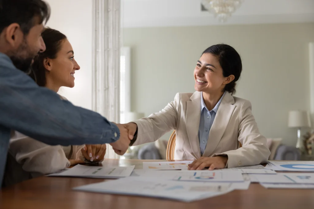 Glad to gain your trust. Happy smiling female financial advisor handshaking with satisfied couple clients after professional consultation finishing budget papers review helping with finance planning